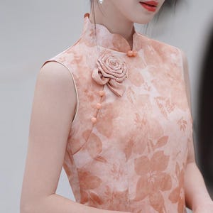 May include: Peach-colored sleeveless dress with a floral pattern. The dress features a mandarin collar, decorative buttons, and a fabric rose detail on the shoulder. The dress is a traditional style, suitable for special occasions.