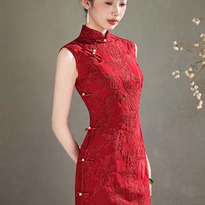 Op de afbeelding: Een mouwloze, rode, bloemen geborduurde jurk met een mandarijnkraag en zijsplitten. De jurk heeft parelachtige knoopsluitingen en een getailleerd silhouet. De stof heeft een subtiele glans en de jurk is een traditionele stijl.