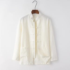May include: Off-white long-sleeve shirt with a mandarin collar and button closures. The shirt has two front pockets and is hanging on a wooden hanger against a white background. The shirt is made of a lightweight fabric.