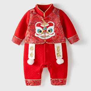 May include: Red baby onesie featuring a lion dance costume design. The onesie has a mandarin collar, long sleeves, and a popper fastening. The lion dance costume is embroidered on the front of the onesie and features a lion's face with gold accents. The onesie also has white tassels with Chinese characters on them.