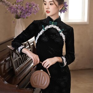 May include: A black, long-sleeved dress with a high collar and floral pattern. The dress features white lace trim and green accents. A brown, round handbag is held in the hand. The background includes a piano and a vase of purple flowers.