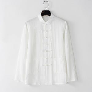 Pode incluir: Uma camisa branca de manga comprida com gola mandarim e fecho de botões. A camisa tem dois bolsos frontais e está pendurada num cabide preto. O tecido parece ser uma mistura leve de linho ou algodão.