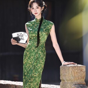 May include: A green, sleeveless cheongsam dress with a floral pattern. The dress has a high collar and a side slit. The person in the image is holding a black and white fan. The dress is a traditional Chinese garment.