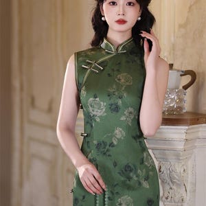 May include: A sleeveless green dress with a floral pattern. The dress has a mandarin collar and frog closures. The model is wearing pearl earrings and holding a beaded necklace. The dress is a traditional Chinese style.