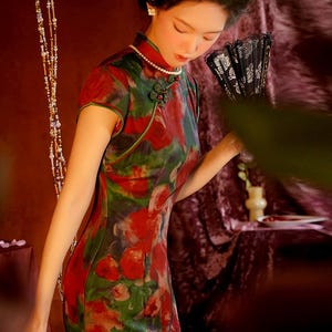 Könnte beinhalten: Eine Frau in einem traditionellen chinesischen Kleid mit einem Blumenmuster in Rot, Grün und Weiß. Das Kleid hat einen Stehkragen und kurze Ärmel. Sie hält einen schwarzen Fächer. Eine Perlenkette und Ohrringe vervollständigen den Look.