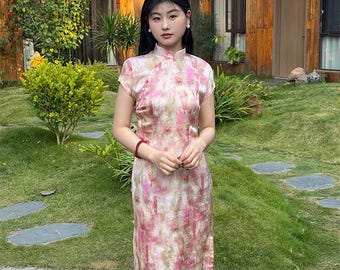 May include: A pink and white floral print dress with a mandarin collar and cap sleeves. The dress is floor-length and has a slim fit. The background includes a wooden building and green foliage.