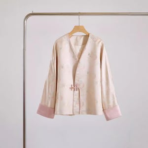 May include: A pale pink jacket with a floral pattern and a decorative button closure. The jacket has long sleeves with pink trim and a V-neck.