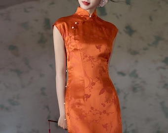 May include: An orange, sleeveless, satin dress with a mandarin collar and floral pattern. The dress features pearl buttons and a side slit. A small, red tassel fan is held in the hand.