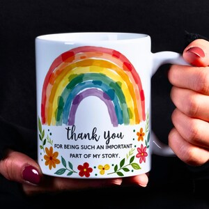 Op de afbeelding: Witte keramische mok met een kleurrijk regenboogontwerp en de tekst "dank je". De mok heeft een boodschap van waardering, met bloemen- en bladaccenten. De mok wordt tegen een zwarte achtergrond gehouden.