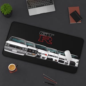 May include: A rectangular black desk mat featuring a graphic illustration of five white Nissan GT-R sports cars lined up in a row. Above the cars, the text GT-R is printed in a bold white and red outline font. The mat has a smooth surface.