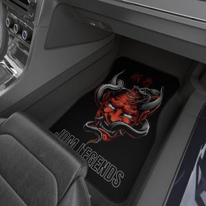 May include: A black car floor mat featuring a red Hannya mask graphic with black horns and a snake coiled around it. The text JDM LEGENDS is printed in white capital letters at the bottom, with red Japanese kanji characters above the mask.