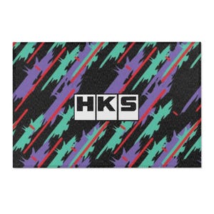 May include: A rectangular mat featuring a black background with a repeating diagonal pattern of teal and purple paint-like brush strokes and thin red lines. The letters HKS are printed in bold black font inside three white square boxes in the center.