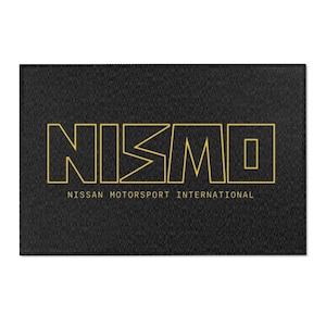 May include: A rectangular black mat featuring the yellow outlined text NISMO in a bold, stylized font. Below the main logo, the text NISSAN MOTORSPORT INTERNATIONAL is printed in a smaller, clean yellow sans-serif typeface on the dark surface.