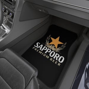 May include: A black car floor mat featuring the Sapporo Premium Beer logo in white text with a gold star and wheat graphic. The design includes the word Sapporo in large white letters, with Premium Beer underneath, positioned in a vehicle interior.