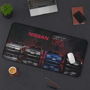 May include: A rectangular desk mat featuring a graphic print of four Nissan GT-R sports cars in silver, grey, blue, and black. The design includes the red text NISSAN and the red outlined GT-R logo set against a dark, smoky background.
