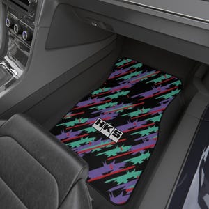 May include: A rectangular car floor mat featuring a black background with a repeating pattern of diagonal teal, purple, and thin red brushstroke-style lines. The center of the mat displays a white rectangular logo with the black text HKS.