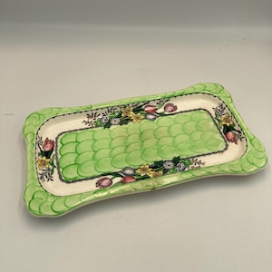 May include: A rectangular ceramic tray with a scalloped edge. The tray features a green, textured interior and a white border adorned with floral patterns in pink, yellow, and blue. The tray's edges are a lighter shade of green.