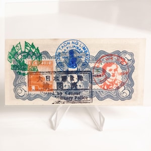 May include: A vintage Philippine banknote, featuring intricate designs and stamps in green, blue, orange, and red. The bill displays text in multiple languages and is held by a clear acrylic stand. The banknote has a denomination of 5.
