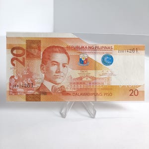 May include: A twenty-peso Philippine banknote, dated 2019, displayed on a clear stand. The note features a portrait of a man, text in Filipino, and the denomination "20" in multiple locations. The note is orange and brown.