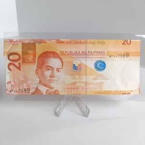 Op de afbeelding: Een Filipijnse bankbiljet van twintig peso, weergegeven in een doorzichtige plastic houder. De biljet toont een portret van een man en de tekst "Republika ng Pilipinas". Het nummer "20" is rechtsboven en linksonder zichtbaar.