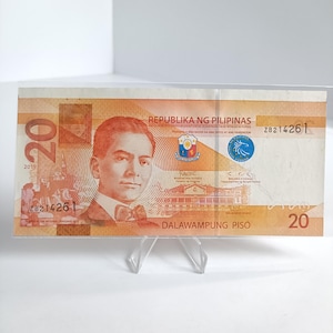 Puede incluir: Un billete filipino de veinte pesos, con un retrato de un hombre y el texto "REPUBLIKA NG PILIPINAS". El billete es naranja y marrón, con el número "20" en un lugar destacado. El billete está sujeto por un soporte de plástico transparente.