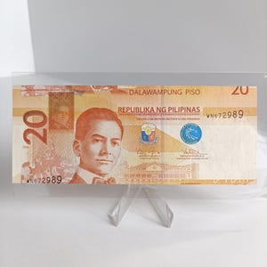 Op de afbeelding: Een biljet van twintig peso uit de Filipijnen, gepresenteerd in een transparante acrylhouder. De biljet toont een portret en de tekst "Republika ng Pilipinas". De biljet is lichtoranje van kleur.