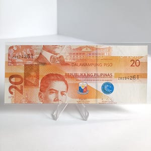 Puede incluir: Un billete filipino de veinte pesos, exhibido en un soporte transparente. El billete es principalmente naranja y presenta retratos, texto y el número 20. El texto incluye "DALAWAMPUNG PISO" y "REPUBLIKA NG PILIPINAS".