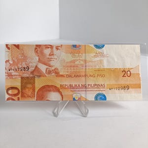 Op de afbeelding: Een Filipijnse bankbiljet van twintig peso, weergegeven in een doorzichtige plastic houder. De biljet toont een portret van een man, de tekst "DALAWAMPUNG PISO" en de woorden "REPUBLIKA NG PILIPINAS". De biljet is voornamelijk oranje en bruin.