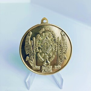 Könnte beinhalten: Ein goldfarbener Metallanhänger mit religiösem Design. Der Anhänger zeigt zwei Engel mit Flügeln, einen Sonnenstrahl und die Worte "EURE SANCTA CORPORA JESU" und "ASSISTIS NOBIS DOMINUS".