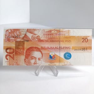 May include: A twenty-peso Philippine banknote, displayed on a clear plastic stand. The note features portraits, text in Tagalog and English, and the number 20. The banknote is orange and brown.