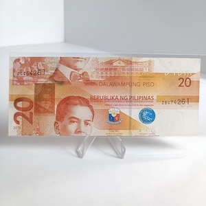 May include: A twenty-peso Philippine banknote, featuring portraits and text. The note displays the text "DALAWAMPUNG PISO" and "REPUBLIKA NG PILIPINAS". The number "20" is prominently displayed. The note is held in a clear plastic holder.
