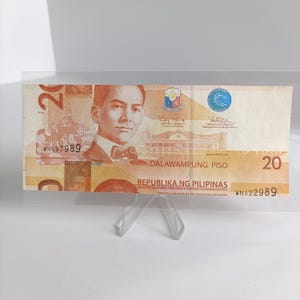 Op de afbeelding: Een Filipijnse bankbiljet van twintig peso, gepresenteerd in een transparante acrylhouder. De biljet toont een portret van een man, de tekst "DALAWAMPUNG PISO" en het getal "20". De bankbiljet is oranje, bruin en wit.
