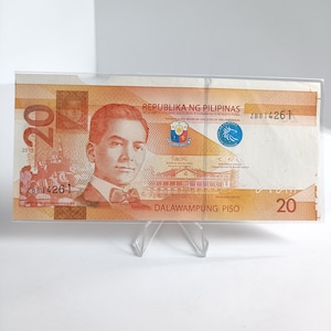 Op de afbeelding: Een Filipijnse bankbiljet van twintig peso, met een portret van een man, de tekst "REPUBLIKA NG PILIPINAS" en het nummer "20". Het bankbiljet wordt weergegeven in een doorzichtige plastic houder op een doorzichtige standaard.