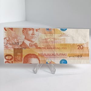 May include: A twenty-peso Philippine banknote displayed in a clear acrylic holder. The bill features a portrait of a man with a bow tie and the text "DALAWAMPUNG PISO". The banknote is predominantly orange and brown.