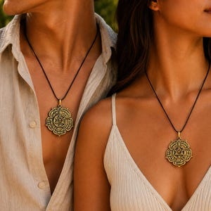 May include: Two people wearing matching gold-colored pendant necklaces with a black cord. The pendants have a detailed, circular design with geometric shapes and text. The person on the left is wearing a light-colored button-down shirt, and the person on the right is wearing a white tank top.
