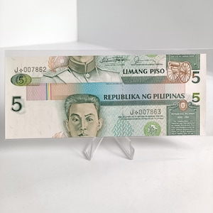Puede incluir: Un billete filipino de cinco pesos, exhibido en un soporte de plástico transparente. El billete presenta retratos de figuras históricas y el texto "LIMANG PISO" y "REPUBLIKA NG PILIPINAS". El billete es verde, azul y rosa.