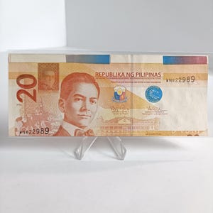 Op de afbeelding: Een Filipijnse bankbiljet van twintig peso, weergegeven in een doorzichtige plastic houder. De biljet toont een portret van een man en de tekst "Republika ng Pilipinas". De bankbiljet wordt vastgehouden door een doorzichtige plastic standaard.