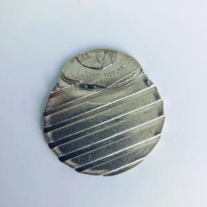 Puede incluir: Una moneda de plata con las palabras "JOSE RIVERA" inscritas en ella. La moneda tiene una superficie texturizada con líneas que la atraviesan.