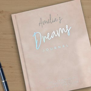 May include: A blush-colored journal with the words "Amelia's Dreams Journal" in silver and light blue script. A dark blue fountain pen rests on a wooden surface next to the journal. The journal is a notebook.