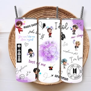 May include: Three white tumblers with purple, pink, and blue designs featuring cartoon characters and text. The text includes song titles and lyrics from the K-pop group BTS. The tumblers are on a wooden surface.