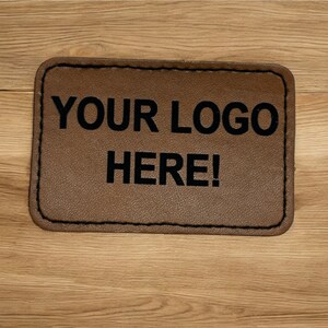 Può includere: Un cerotto in pelle marrone con cuciture nere attorno ai bordi. Il testo "YOUR LOGO HERE!" è stampato in nero sul cerotto.