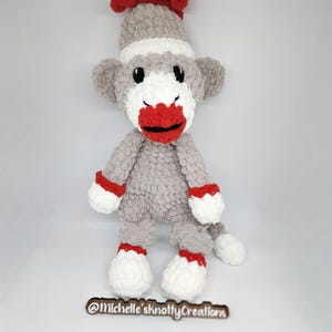 May include: A handmade gray sock monkey with a red tuft of hair, red lips, and white accents. The monkey has black eyes and red details on its hands and feet. The product is a plush toy.