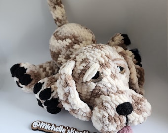 May include: A crocheted dog toy with brown and white fur, black paw pads, and a pink tongue sticking out. The toy is lying on its side with its head turned to the side. The toy is made by Michelle's Knotty Creations.