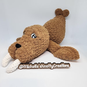May include: A brown crocheted walrus plush toy with white tusks, black eyes, and a black nose. The plush is lying on a white surface. The text "@Michelle'sknottyCreations" is below the plush.