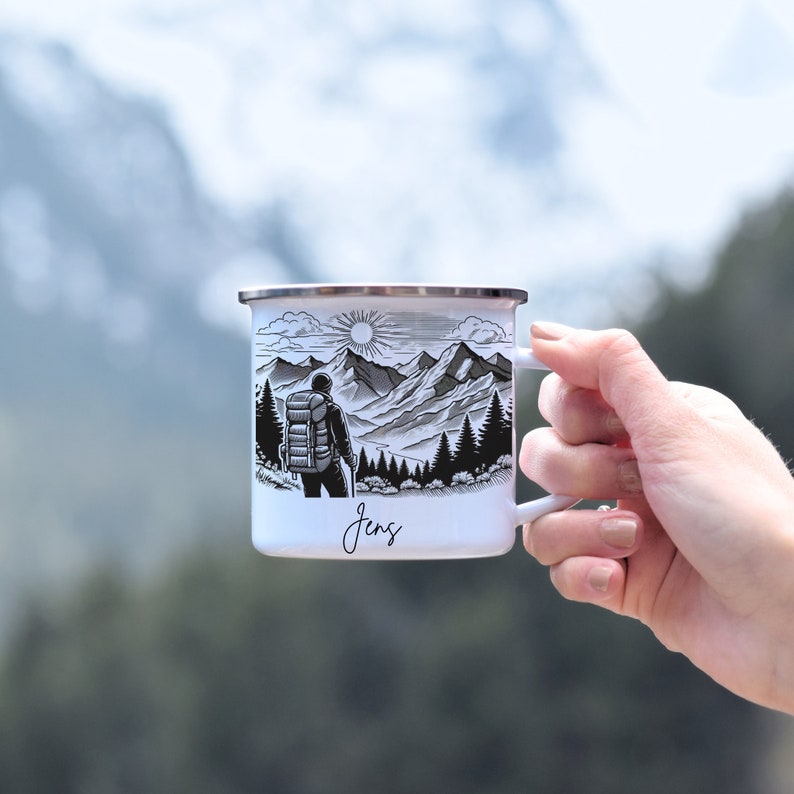 May include: White enamel camping mug with a black and white illustration of a hiker with a backpack in front of a mountain range. The mug has a black rim and handle and the name "Jens" is written in cursive at the bottom.