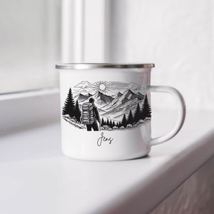 May include: White enamel mug with a black and white illustration of a person hiking in the mountains. The mug has a silver rim and handle. The text "Jens" is printed on the bottom of the mug.