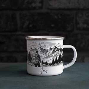 May include: White enamel camping mug with a black and white illustration of a hiker in a mountain landscape. The mug has a silver rim and a black handle. The name "Jens" is written on the bottom of the mug.