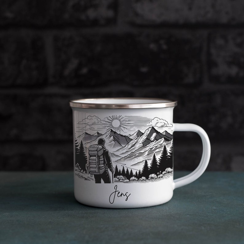 May include: White enamel camping mug with a black and white illustration of a hiker in a mountain landscape. The mug has a silver rim and a black handle. The name "Jens" is written on the bottom of the mug.