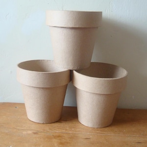 May include: Three stacked, beige-colored flower pots. Each pot has a wide, rounded rim and a cylindrical body. The pots are made of a light-colored, textured material, possibly paper or cardboard, and are arranged on a wooden surface.