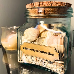 May include: A square, light blue-tinted glass jar with a cork lid, filled with various white and tan seashells and small fragments. A rectangular gold-toned label on the front features the black printed text, Kimberly's Collection.